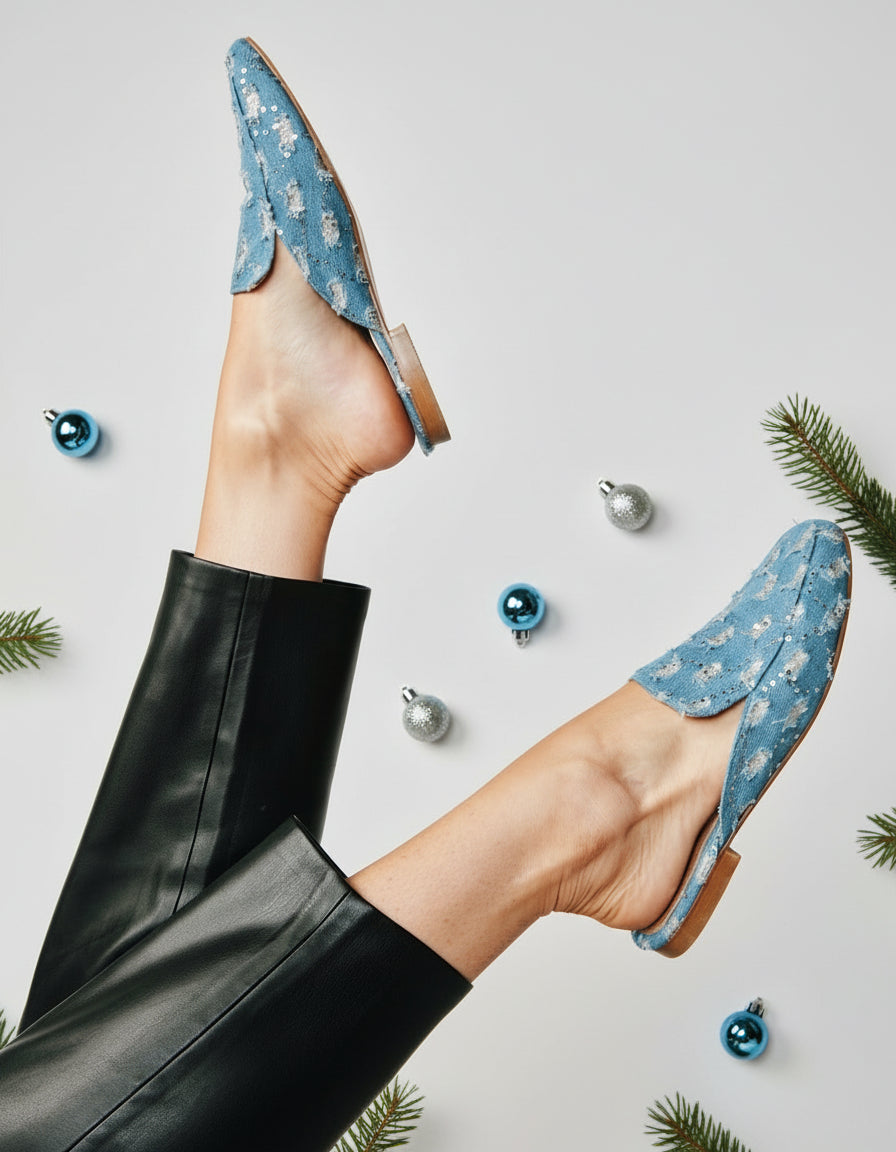A pair of light blue denim mules with a pointed toe and flat sole.