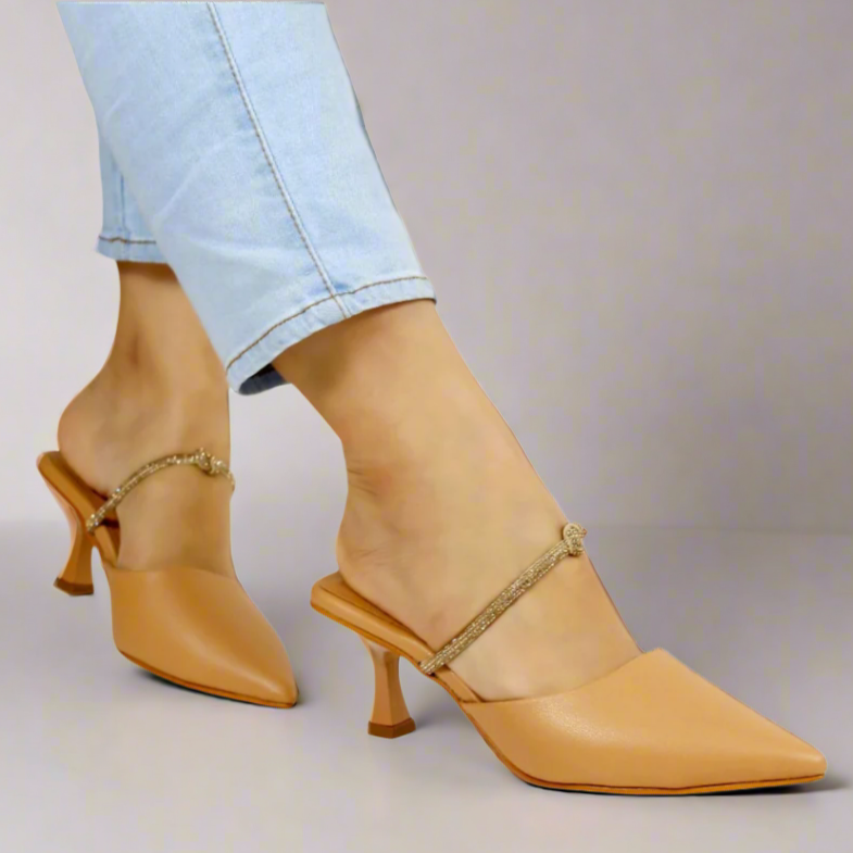 A pair of beige pointed-toe mules with kitten heels, displayed on a person wearing light blue jeans. The shoes have a strap with a buckle across the instep.