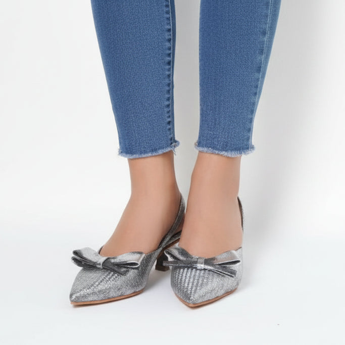 A pair of silver metallic kitten heels with a low and slim heel, featuring a functional strap and a bow detail on the front.