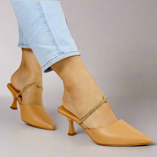 A pair of beige pointed-toe mules with kitten heels, displayed on a person wearing light blue jeans. The shoes have a strap with a buckle across the instep.